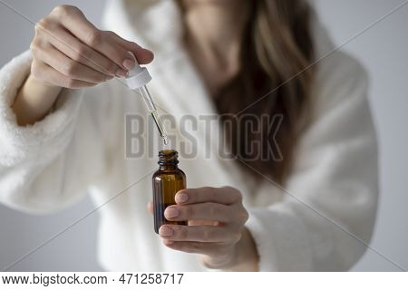 Young Woman In A Bathrobe Is Holding A Bottle With A Dropper Of Oil Or Serum For Face And Body Care.