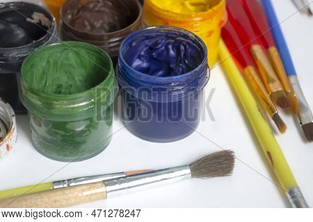 Jars Of Colored Paint Stand Next To The Brushes. Watercolor And Oil Paints On A White Table. For Art