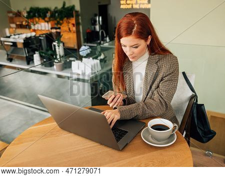 Young Female Entrepreneur Working In A Coffee Shop. Woman With Ginger Hair Typing On A Laptop And Ho