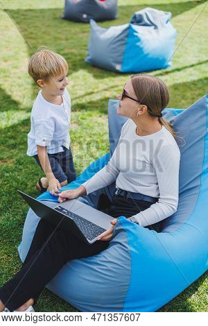 A Mother With Her Son On A Green Lawn, A Mother On Maternity Leave Is Working On A Laptop In The Par
