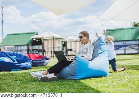 Mother With Laptop, Her Son Playing Outdoors On Green Lawn. Rest And Work With A Child In The Park