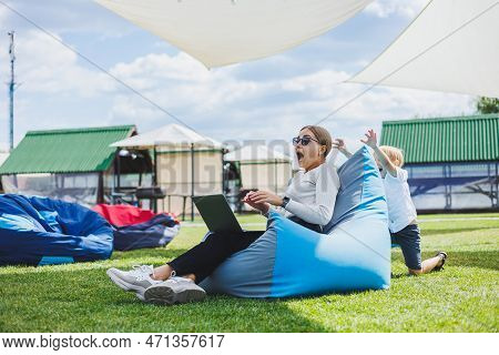 Mother With Laptop, Her Son Playing Outdoors On Green Lawn. Rest And Work With A Child In The Park