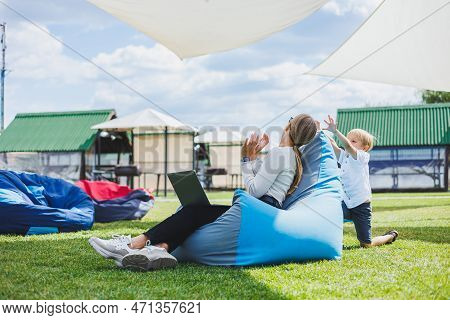 Mother With Laptop, Her Son Playing Outdoors On Green Lawn. Rest And Work With A Child In The Park