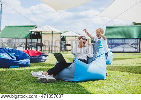 Mother With Laptop, Her Son Playing Outdoors On Green Lawn. Rest And Work With A Child In The Park