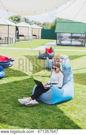 Mother With Laptop, Her Son Playing Outdoors On Green Lawn. Rest And Work With A Child In The Park
