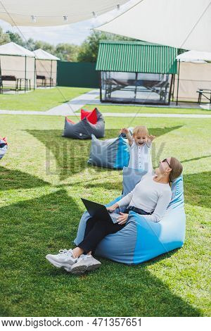 Mother With Laptop, Her Son Playing Outdoors On Green Lawn. Rest And Work With A Child In The Park