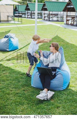 Mother With Laptop, Her Son Playing Outdoors On Green Lawn. Rest And Work With A Child In The Park
