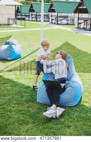 Mother With Laptop, Her Son Playing Outdoors On Green Lawn. Rest And Work With A Child In The Park