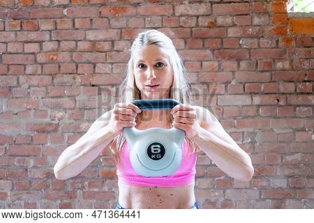 Blonde Young Muscular Woman With Sporty Looks Doing High Pull Exercise With Kettlebells On Hard Trai
