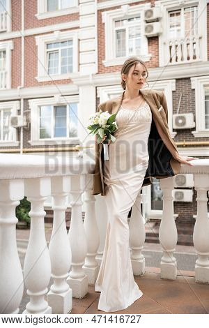 Full Length Of Young Bride In Wedding Dress And Beige Blazer Holding Bouquet Near House.