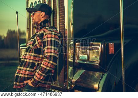 Closeup Of Caucasian Trucker Leaning On His Semi Truck Grille While Taking Rest During Road. Trucker