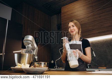 Beautiful Female Confectioner Dressed In Apron Beats Cream With Mixer In Modern Kitchen