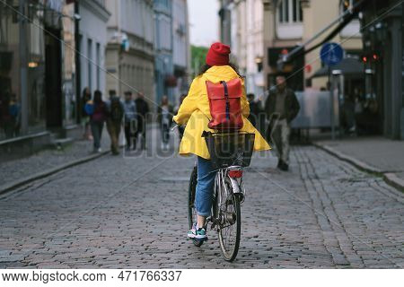 A Young Woman In A Yellow Windbreaker And A Red Winter Hat Rides A Bicycle On A Paved Pedestrian Pat