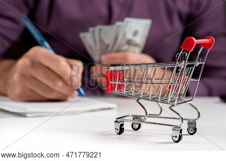 Man Calculates The Budget. Shopping Cart A On Table With Calculator And Paper And Mohey In Hadns. Bu