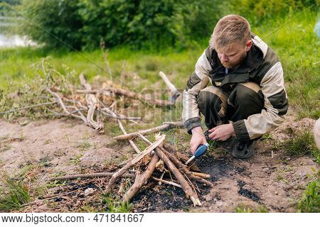 Bearded Survivalist Male In Raincoat Starting Fire Using Gas Lighter On Bank Of River For Cooking An