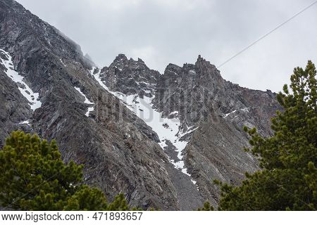 Gloomy Mountain Landscape With High Sharp Rockies With Snow In Gray Low Clouds. Closeup Of Sharp Roc