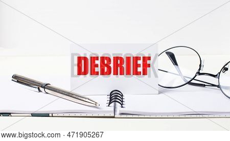 Close-up Of A White Card With The Text Debrief, A Stylish Pen And Glasses On A Desktop.