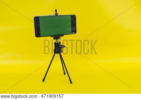 Black Mini Tripod With Steel Case, Ballhead And Smartphone With Greenscreen Isolated On Yellow Backg