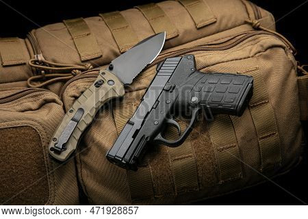 A Small Modern Pistol And Penknife On A Brown Military Backpack. Self Defense Kit. Dark Background.