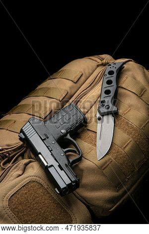 A Small Modern Pistol And Penknife On A Brown Military Backpack. Self Defense Kit. Dark Background.