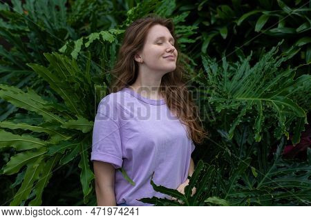 Portrait Of Young Woman Standing On An Ivy, Fern Wall Or In Greenery, Breathing, Smiling And Looking