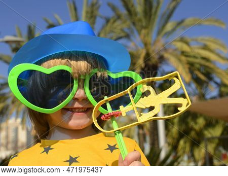 Carnival, Purim In Israel. A Cheerful Little Boy In A Hat, Funny Glasses, A Wig With A Rattle, Raash