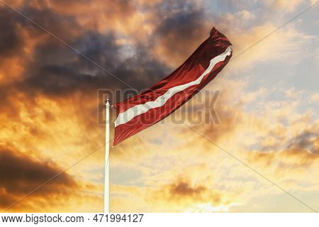 The National Flag Of Latvia Flutters In The Wind. Latvian Flag On The Background Of A Cloudy, Orange