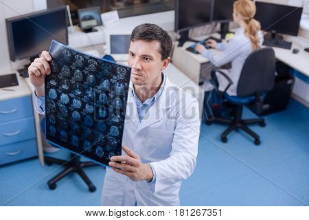 Medical research. Serious experienced male oncologist holding an X ray image and examining it while being in the staffroom