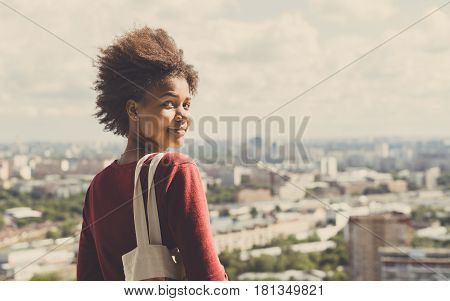 Portrait of young beautiful cute mixed Brazilian teen girl with curly afro hair standing on observation point very high and half-turned to camera sunny summer cityscape below in blurred background