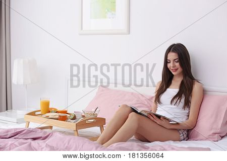 Beautiful young woman having breakfast and reading magazine while sitting on bed at home