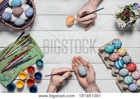 Preparation for Easter. Human hands paint Easter eggs with brushes and paints on an old white wooden background. Top view. Flat lay style