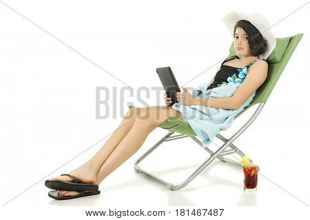 A young teen in beach garb looking up at the viewer from her electronic device with has a tall glass of iced tea by her side.  On white background.