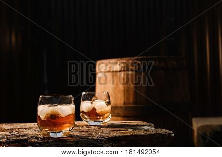 A glass of whiskey with ice on a wooden background close-up in the background of a barrel of whiskey