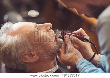Accurate in details .Accurate attentive handsome barberstanding at the workplace and cutting beard of the aged man while using razor