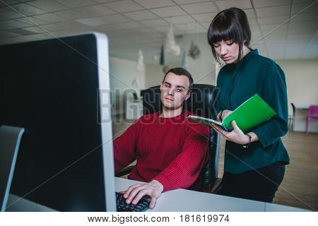 Young office workers. A young man sits at the computer in his chair and young business woman standing next to him and show something in a notebook.