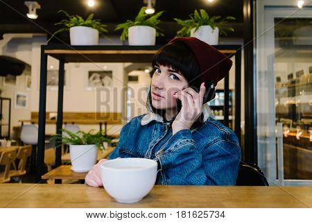 Young happy hipster girl in a cap and a blue denim jacket talking on the phone and drinking a hot drink in a nice cafe
