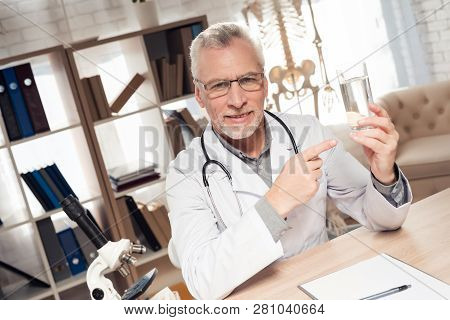 Doctor In White Robe Sitting At Desk In Office With Microscope And Stethoscope. Man Is Holding Water