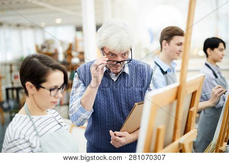 Portrait Of Mature Art Teacher Criticizing Work Of Student Painting Picture On Easel In Art Class, C