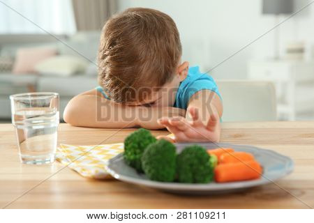 Unhappy Little Boy Refusing To Eat Vegetables At Table In Room