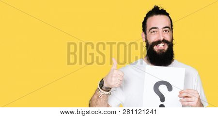 Young man with long hair and bear holding a paper with question mark happy with big smile doing ok sign, thumb up with fingers, excellent sign