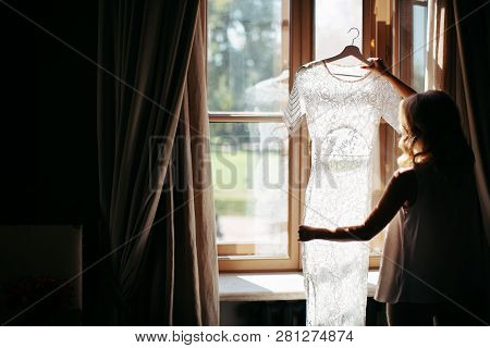 Blonde Woman With Curly Hair Holding And Looking At Beautiful White Wedding Dress On A Hanger, Stand