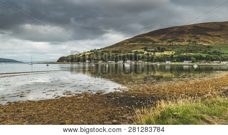 Panoramic view of the Arran island bay. Scotland. Autumn landscape. Scottish shoreline under the grey cloudy sky before the rain. Peaceful countryside scene. Settlement among the wild English nature.