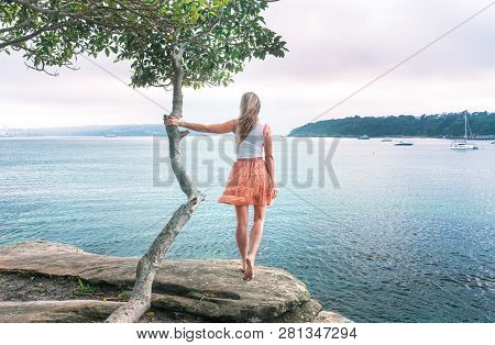 Girl Standing On Rock Ledge Looking Out To Sea Holding On To Lone Tree, The Wind Catching Her Hair A
