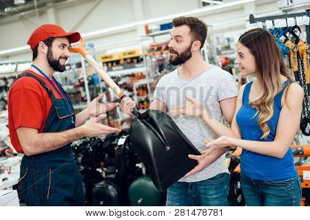 Salesman In Red Shirt And Baseball Cap Is Showing Couple Of Clients New Showel In Power Tools Store.