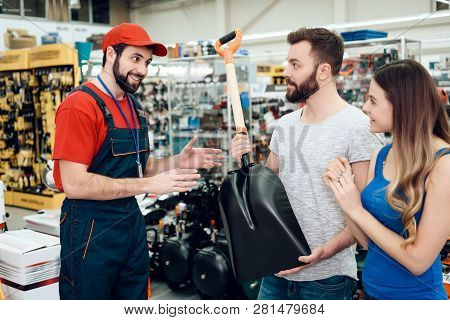 Salesman In Red Shirt And Baseball Cap Is Showing Couple Of Clients New Showel In Power Tools Store.