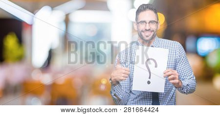 Handsome young business man holding paper with question mark over isolated background happy with big smile doing ok sign, thumb up with fingers, excellent sign