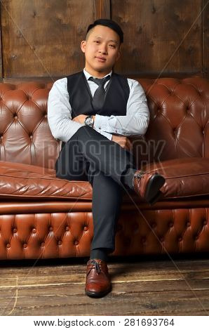 A Successful Asian Man Sits On A Leather Couch, Putting His Foot On His Leg And Crossing His Hands