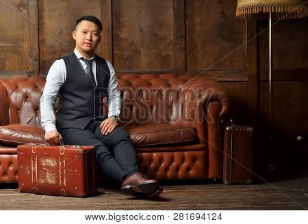 A Young Man In A Suit Sits On A Leather Brown Sofa With A Suitcase