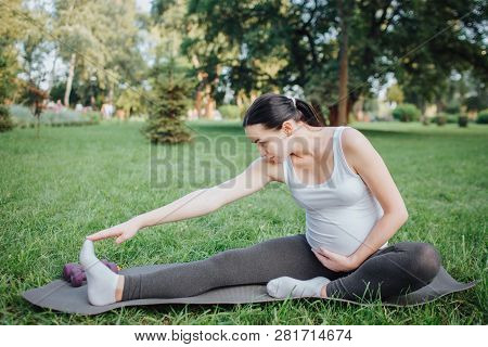 Young Pregnant Woman Stretching Outisde In Park. She Reach Feet With One Hand On Yoga Mate. Model Ke
