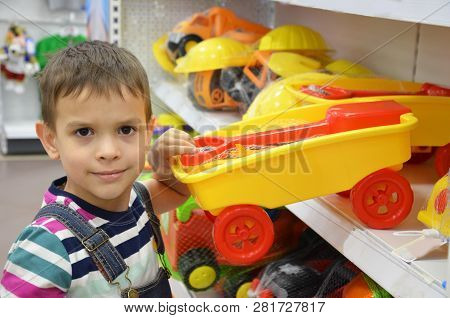 The Boy In Shop With The Toy Machine In Hands. Boy Near The Window With Toys, Big Machine. Chooses A